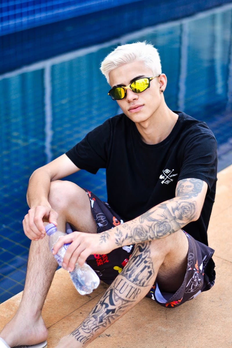 Stylish young man with sunglasses and tattoos sitting poolside in summer fashion.